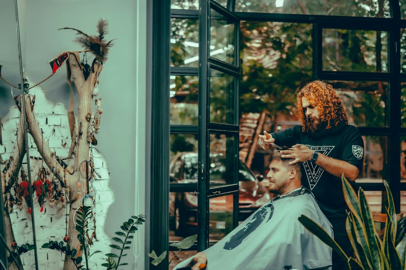 A red-haired barber trims a client's hair with scissors by an open window